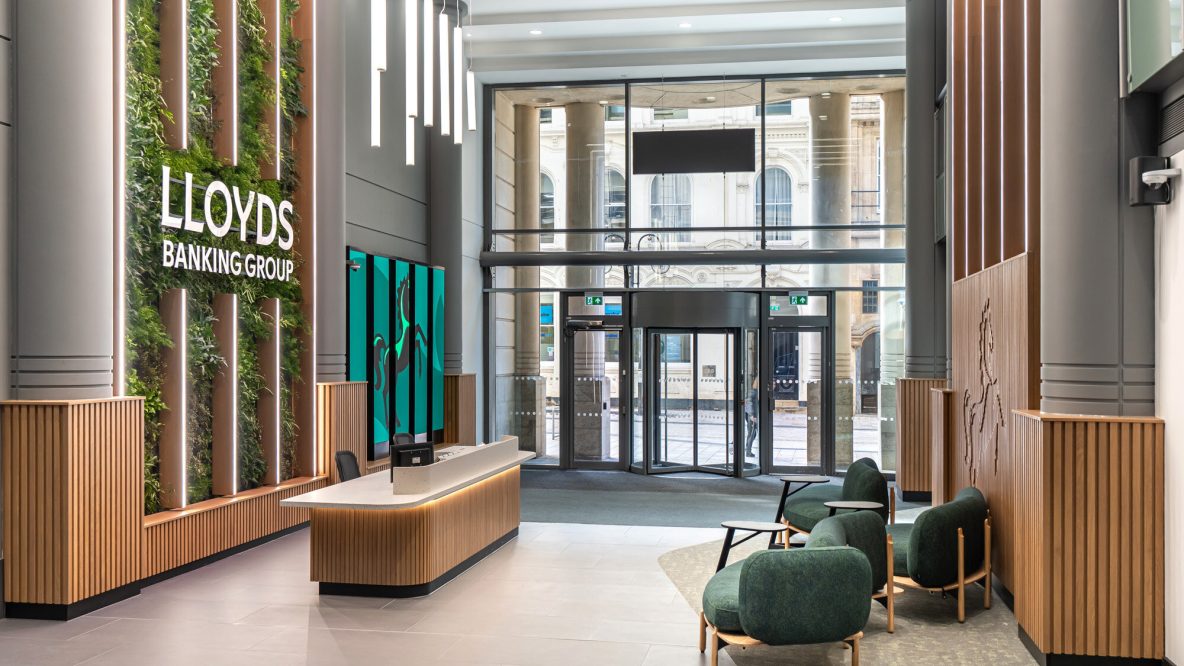 Modern reception area with a vertical garden wall featuring the "Lloyds Banking Group" logo, a white reception desk with wood paneling, green lounge chairs, and a revolving glass door entrance in the background.