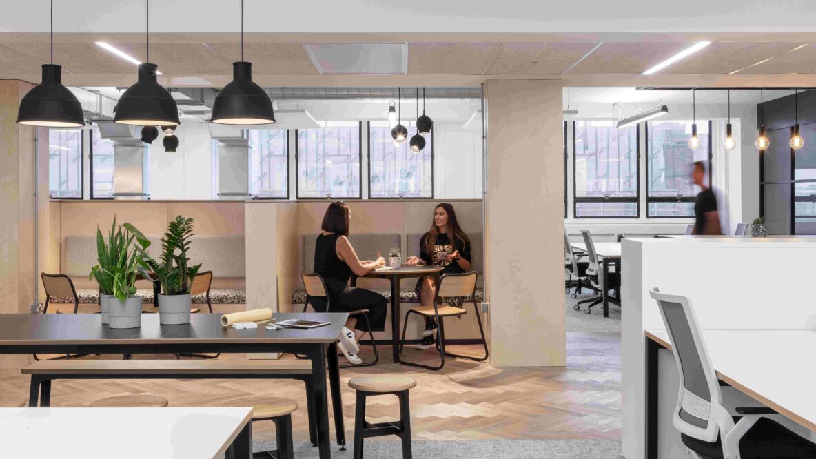 Open plan office space, with long table to the left, two employees sitting in the middle and desk space to the right.