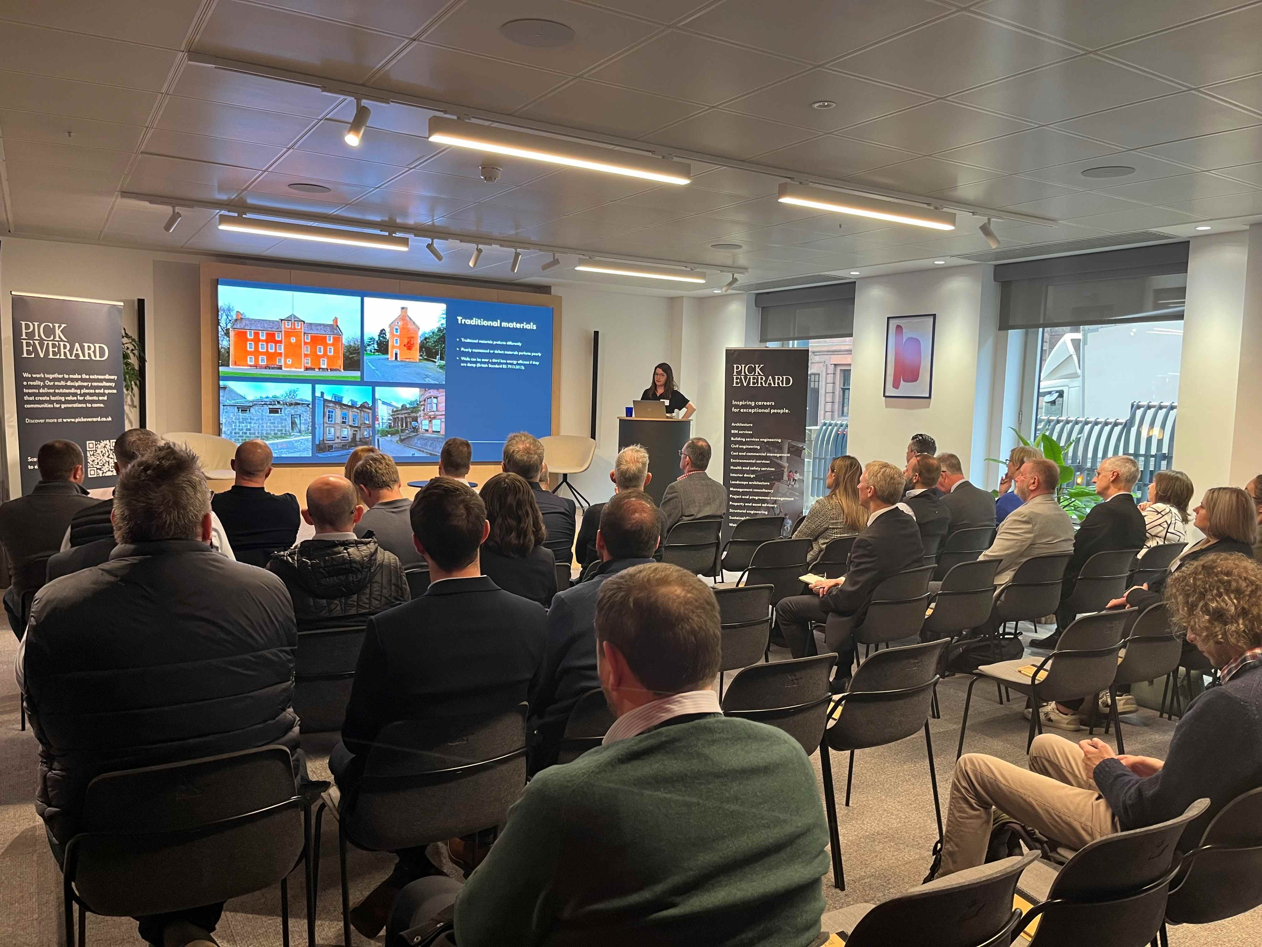 A group of attendees sits in a seminar room, facing a speaker and a presentation showcasing various building designs and materials.