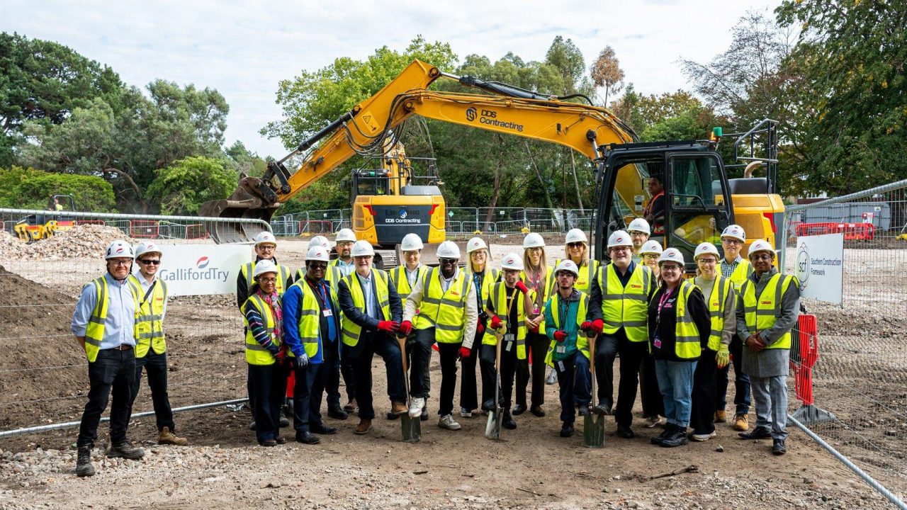 A group of people wearing yellow vests stand at a construction site, facing forward. A few people have spades in their hands.