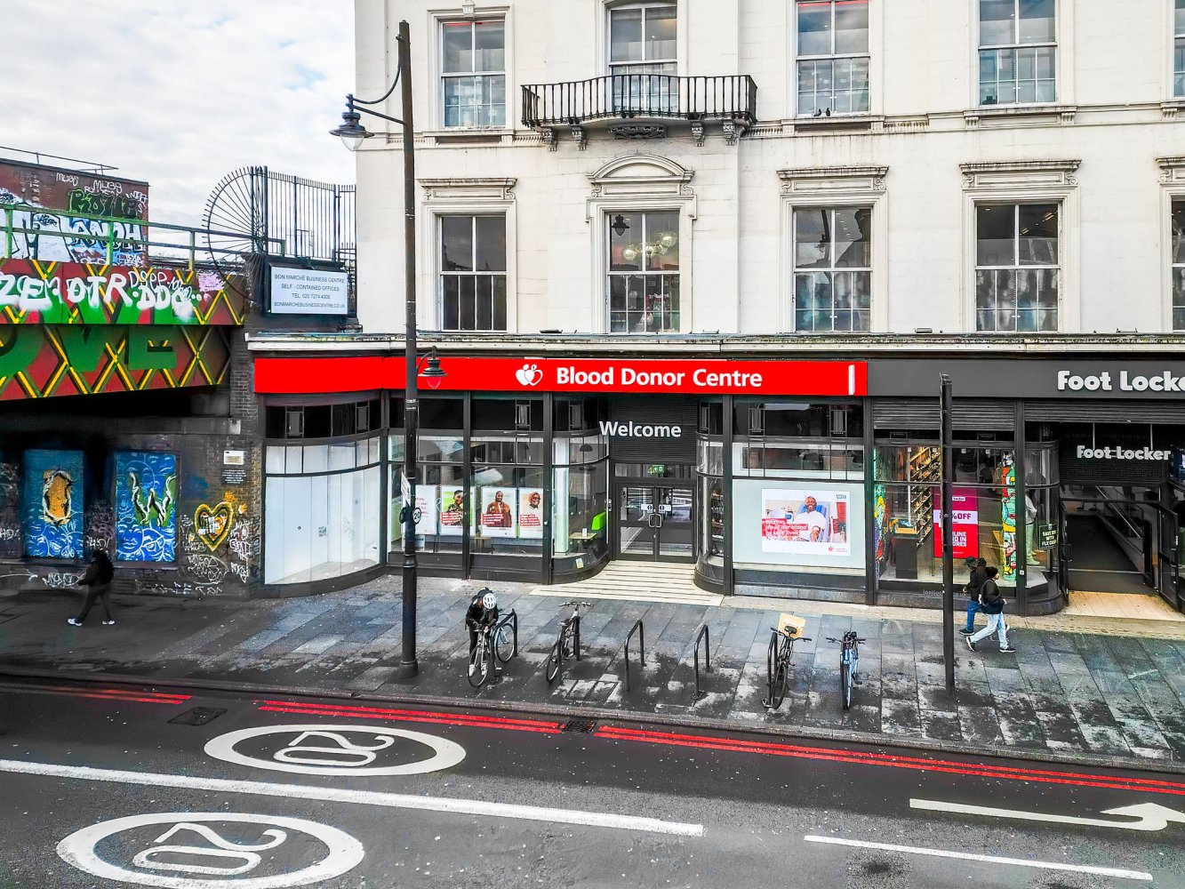 Street view of a Blood Donor Centre entrance next to a Foot Locker store. Bicycles are parked outside, and there's graffiti on a nearby wall under a railway bridge.