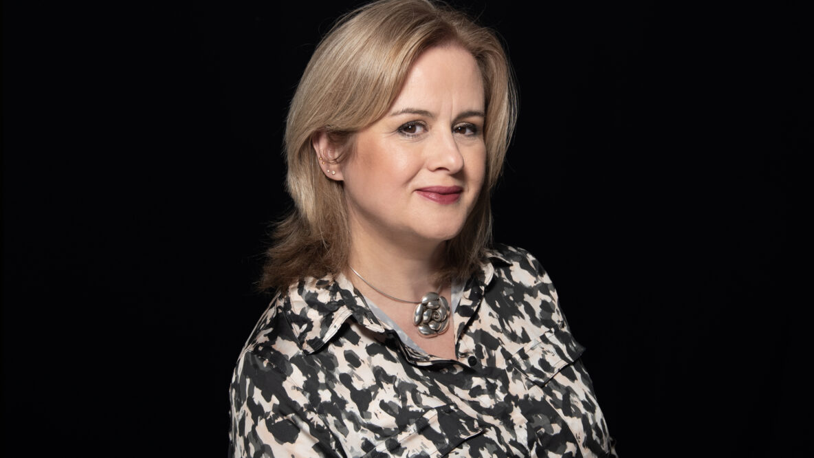 Nicola Hamill smiles, wearing a patterned blouse and a silver necklace against a black backdrop.