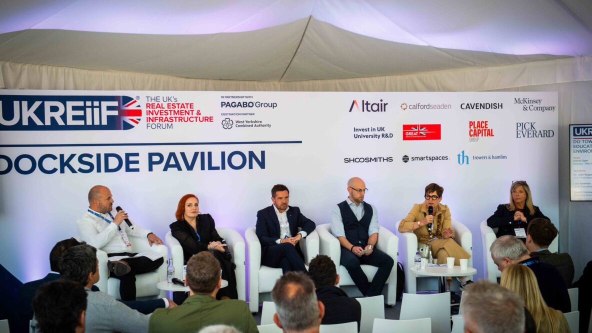 Six panellists sitting on chairs at the front of a room delivering a talk