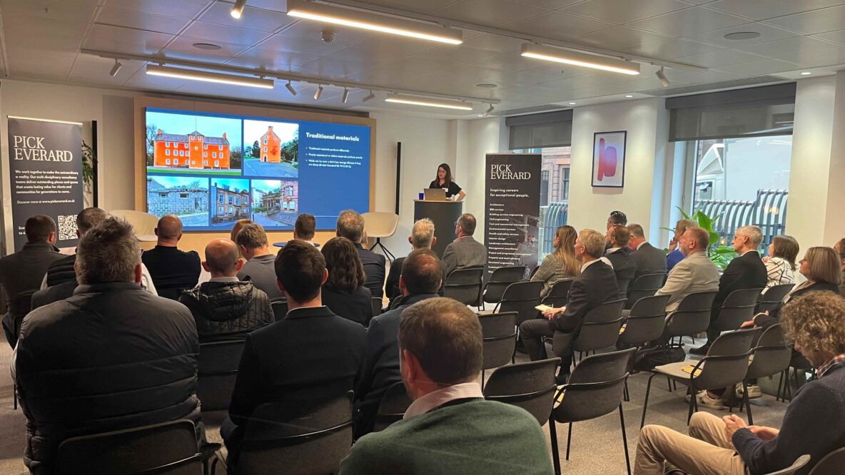 A group of attendees sits in a seminar room, facing a speaker and a presentation showcasing various building designs and materials.