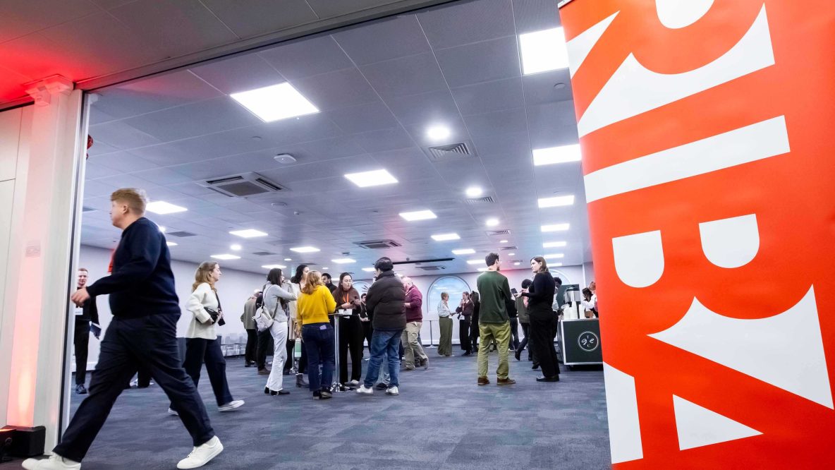 A group of people mingle in a brightly lit conference hall with a large orange banner on the right featuring white text.