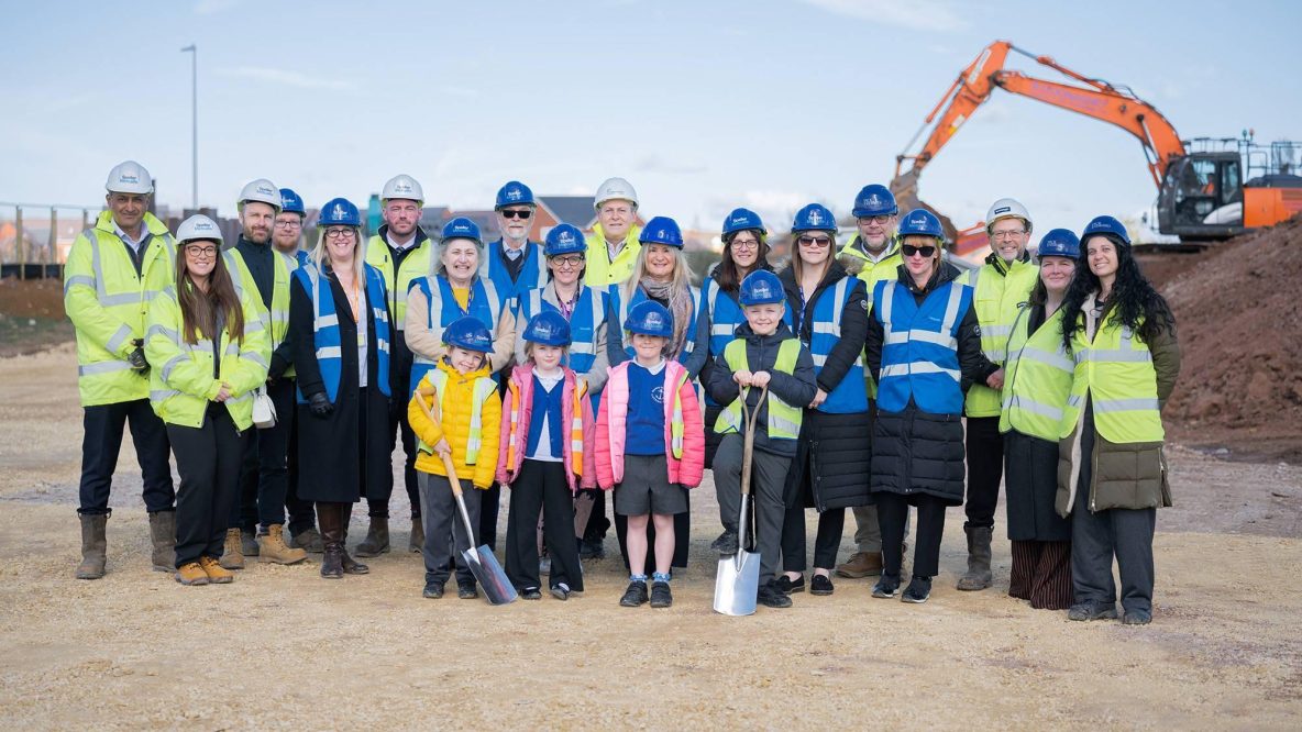 Group of project team and children gathered on site wearing high vis and hard hats smiling at the camera