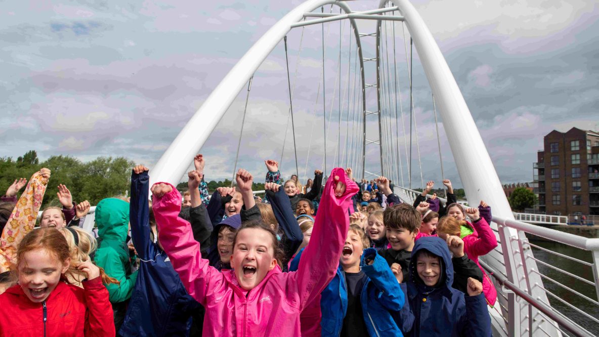 A large group of excited children wearing brightly colored raincoats cheer with their fists raised on a white arched pedestrian bridge.
