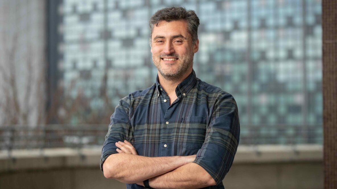 Ricardo smiling in front of a blurred building in the background with his arms crossed